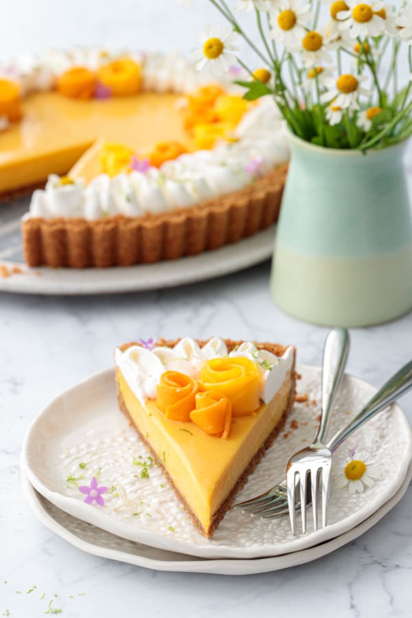 Slice of Mango Lime Tart on a white plate with forks, vase of chamomile flowers and the rest of the tart in the background.