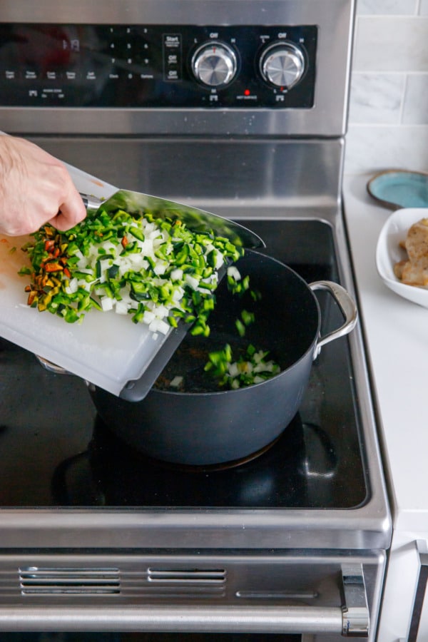Scrapping finely chopped poblano peppers and onions into a saucepan on the stove.