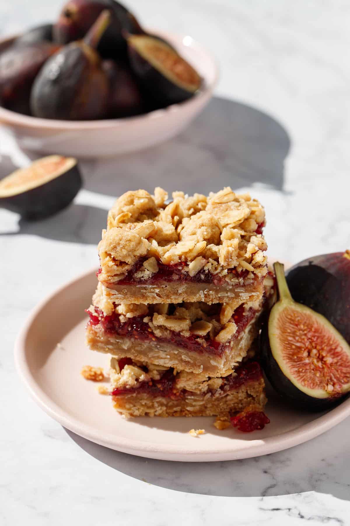 Stack of three Fig, Apple & Vanilla Crumb Bars squares on a pink plate, with fresh figs and a bowl of figs in the background.