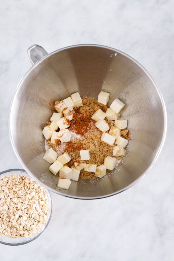Overhead, bowl with flour, brown sugar, spices, and cubes of softened butter sprinkled on top; bowl of oats on the side.