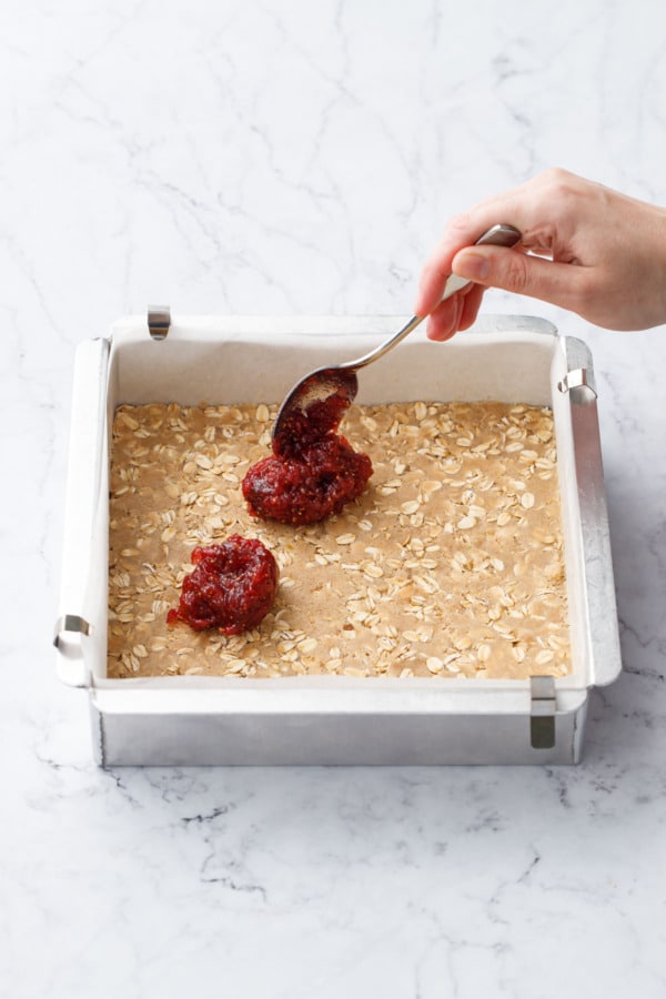 Spoon dropping dollops of deep purple fig apple jam on top of base layer in a square baking pan.