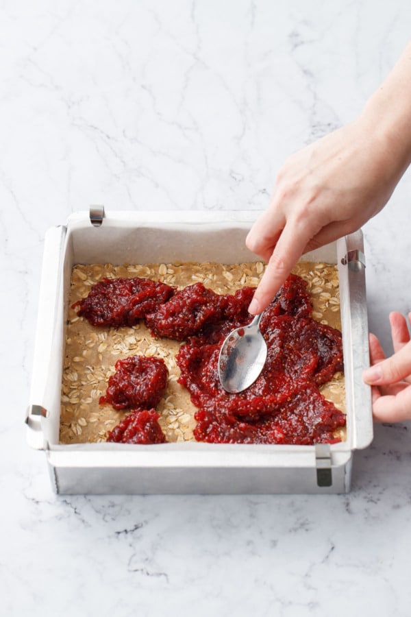 Spoon spreading the deep red jam into an even layer on top of the bottom layer of dough.