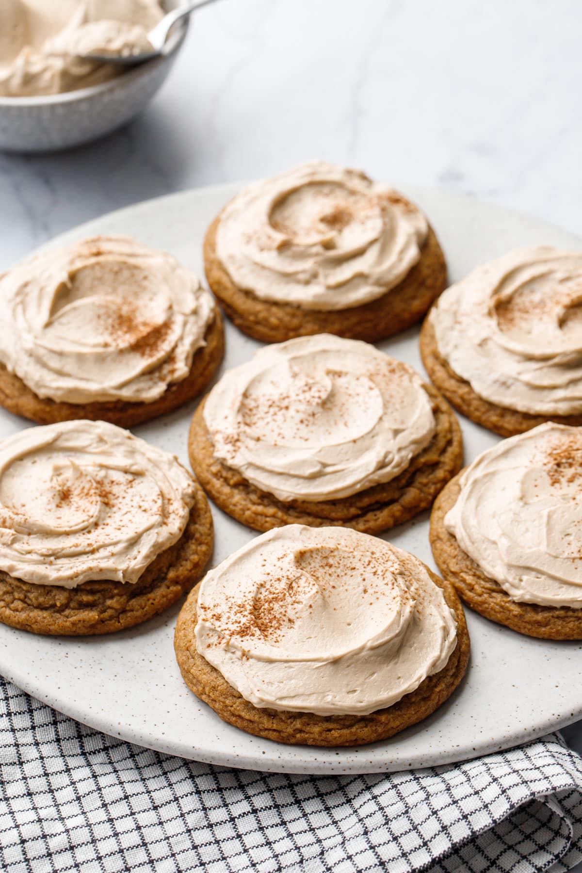 Plate with Caramel Pumpkin Cookies, striped napkin, and a bowl of Salted Caramel Meringue Buttercream in the background.