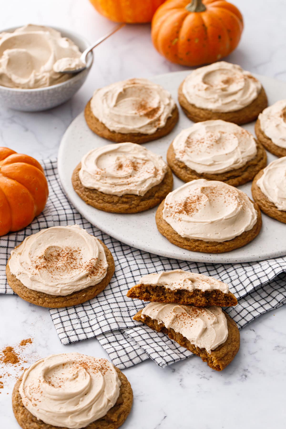 Plate of Caramel Pumpkin Cookies with Salted Caramel Frosting, one cookie broken in half to show the chewy texture inside, with a few small pumpkins in the background.