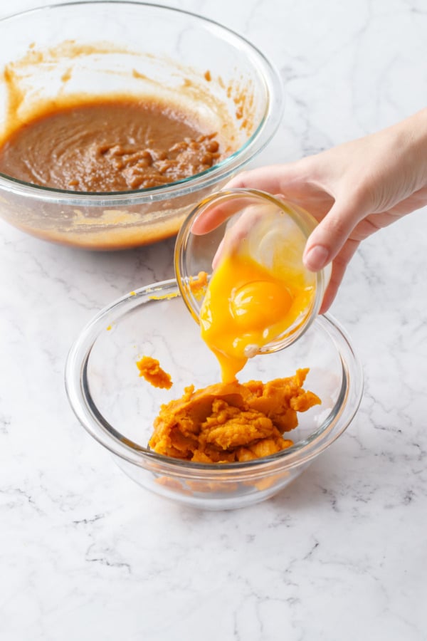 Pouring egg yolks into mixing bowl with pumpkin puree, another bowl of batter in the background.