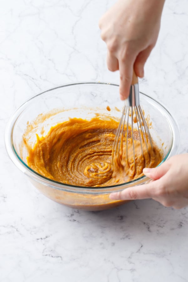 Whisking orange pumpkin cookie dough mixture in a glass mixing bowl with a wire whisk.