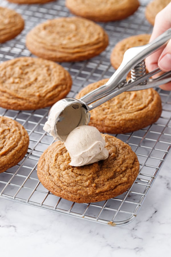 Small cookie scoop dropping a ball of caramel frosting on top of pumpkin cookie on a wire rack.