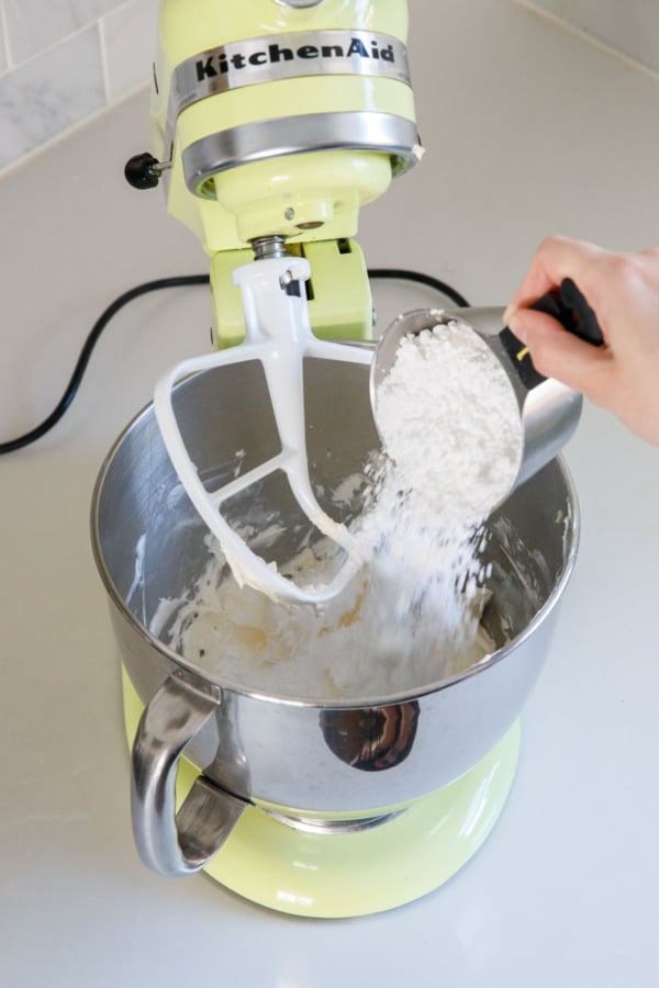 Adding 1 cup of powdered sugar to a stand mixer with meringue frosting in the bowl and paddle attachment.