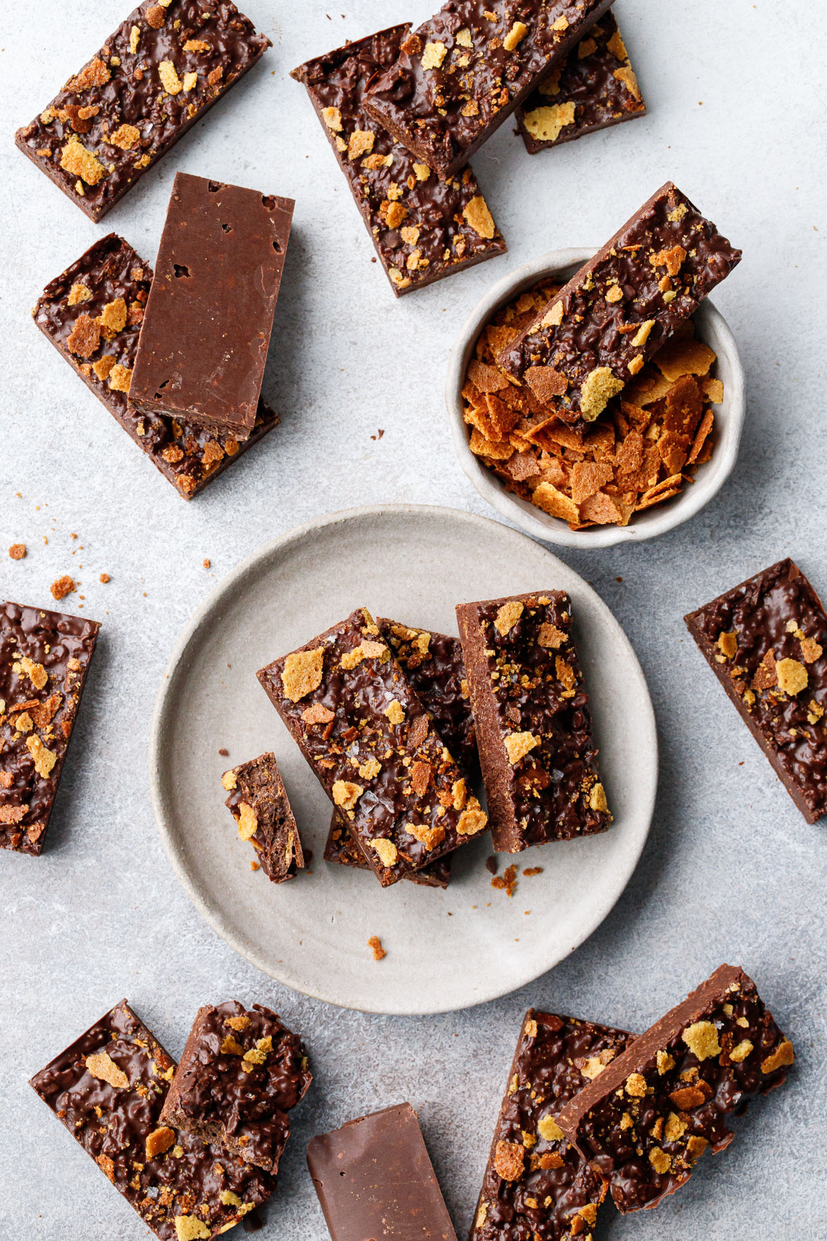 Overhead, bars of Nutella Chocolate Crunch Candy Bark messily arranged on a gray background, a few bars on a plate, and a bowl of feuilletine too.