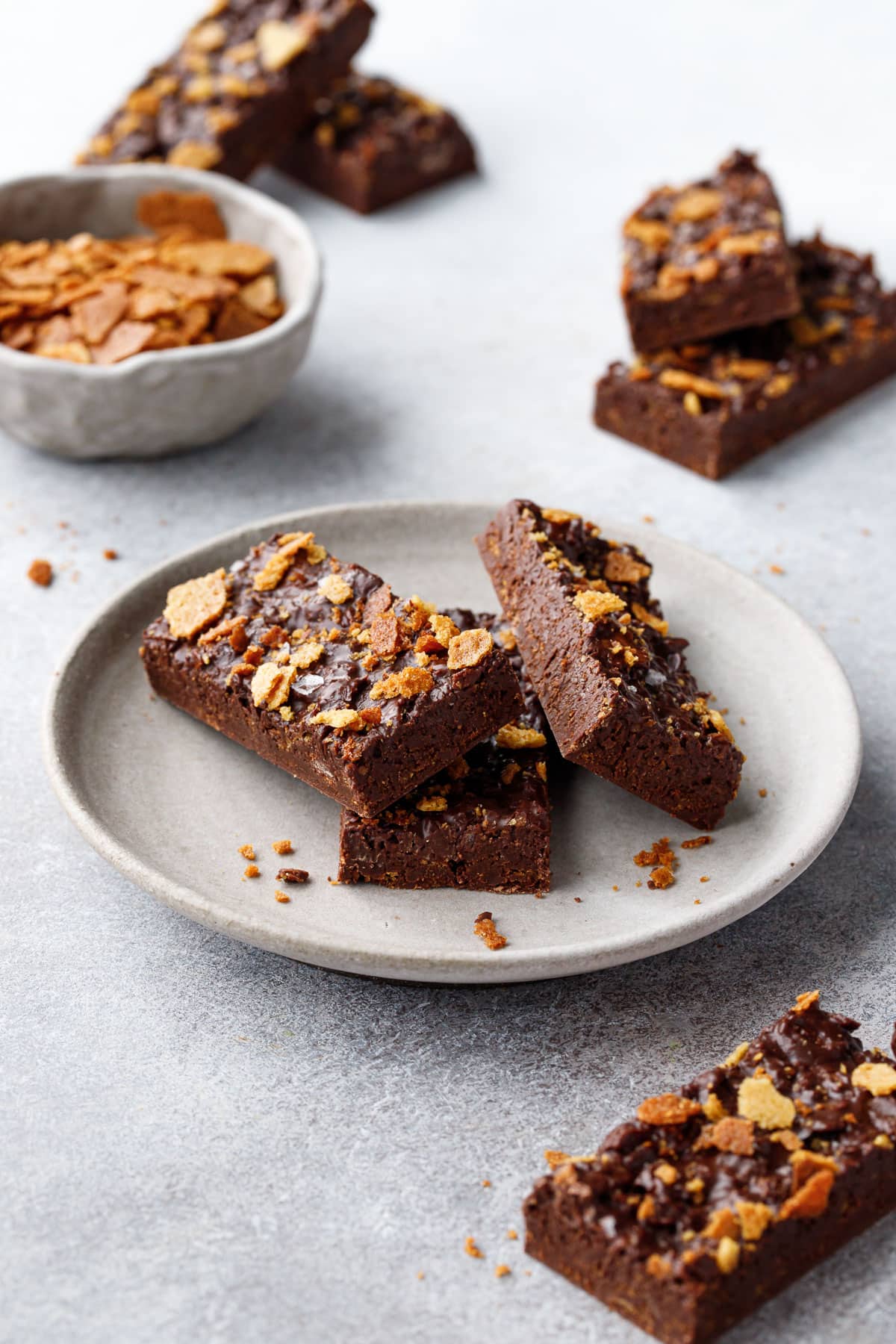Rectangular cut bars of Nutella Chocolate Crunch scattered on a gray background, some piled on a plate and some on the side, with a small bowl of feuilletine in the background.