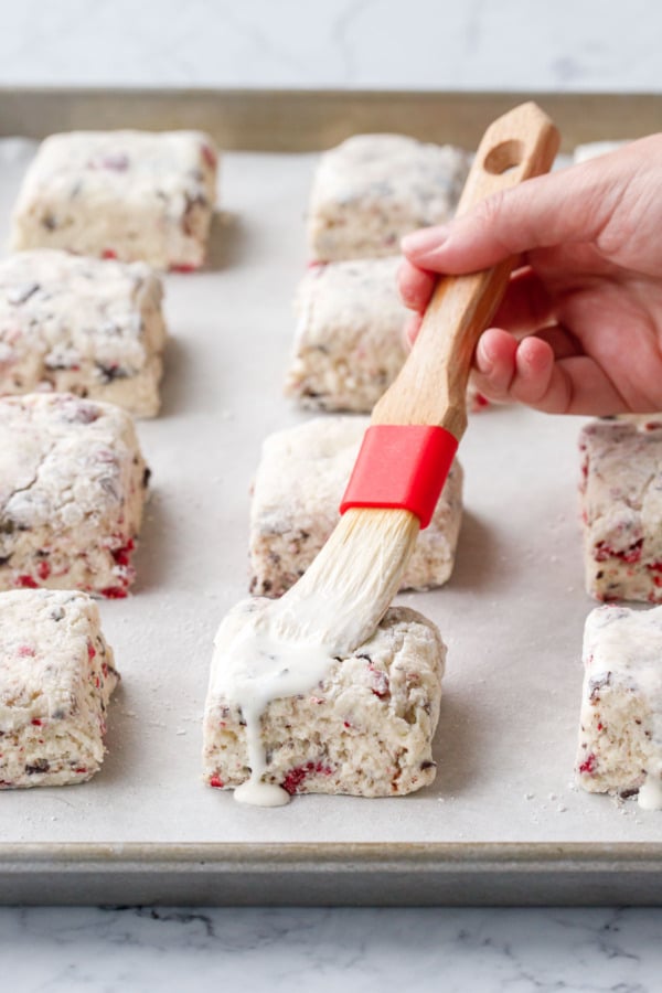 Pastry brush brushing heavy cream onto tops of unbaked scones arranged in rows on a parchment-lined baking sheet.