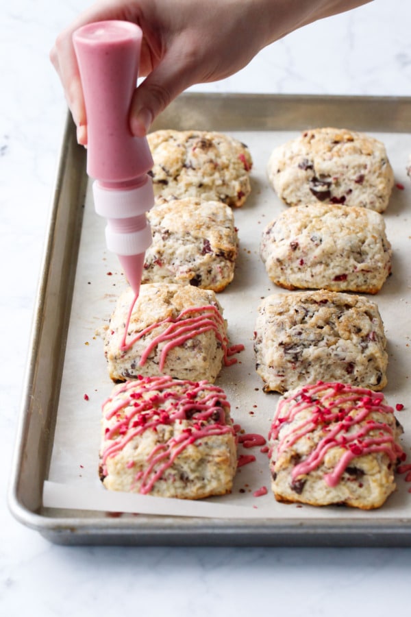 Squeeze bottle filled with raspberry glaze drizzling lines of hot pink raspberry glaze on top of baked scones.