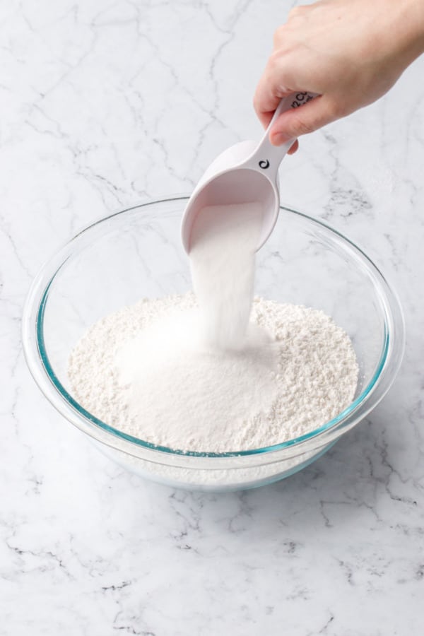 Pouring 1/2 cup of sugar into a mixing bowl along with other dry ingredients.
