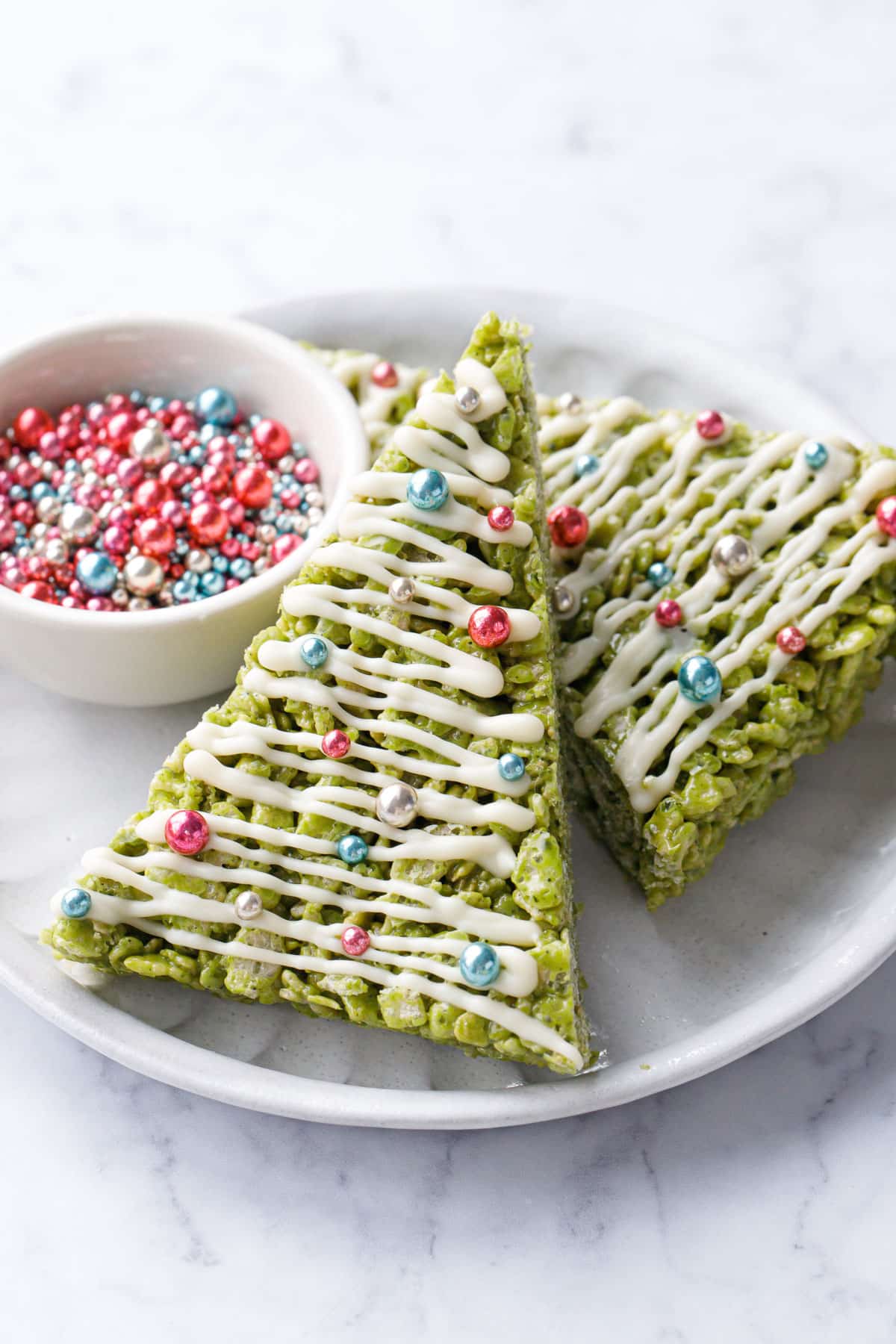 Two green Matcha Rice Crispy Treats decorated to look like Christmas trees on a plate with a bowl of metallic sprinkles.