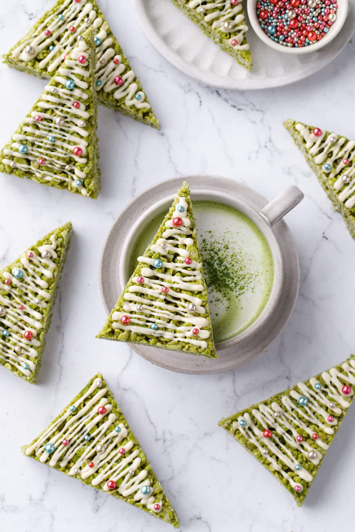 Overhead, Christmas-tree shaped Matcha Rice Crispy Treats arranged randomly on a marble background, one sitting on top of a mug of matcha, plus a bowl of metallic sprinkles.