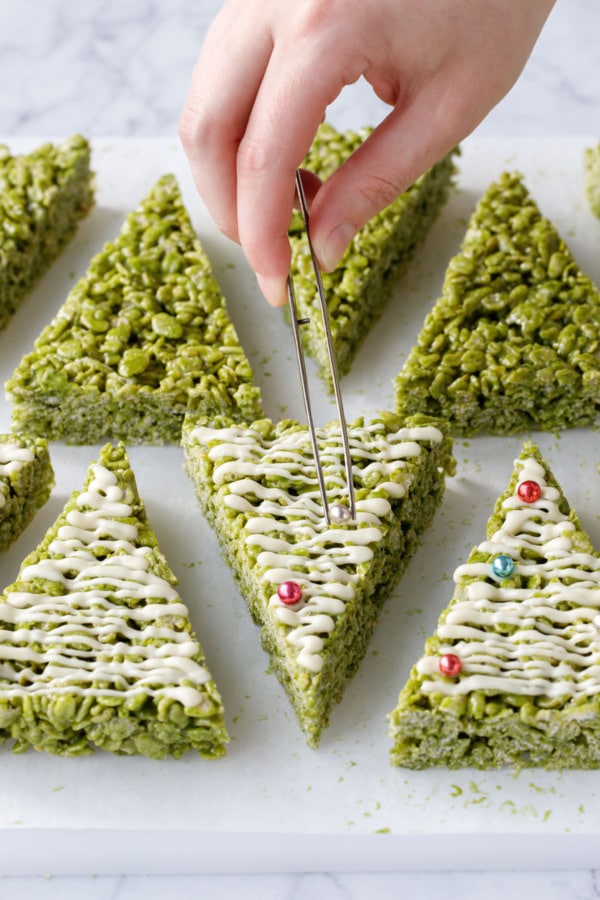 Tweezers placing metallic ball sprinkles that look like ornaments on the green tree-shaped treats.