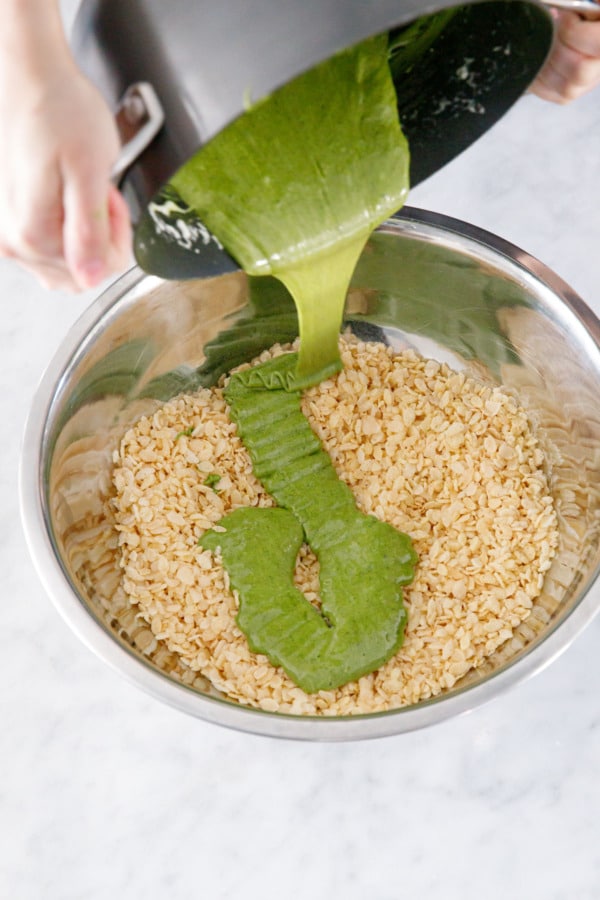 Pouring matcha marshmallow mixture over top of rice crispy cereal in a large metal bowl.