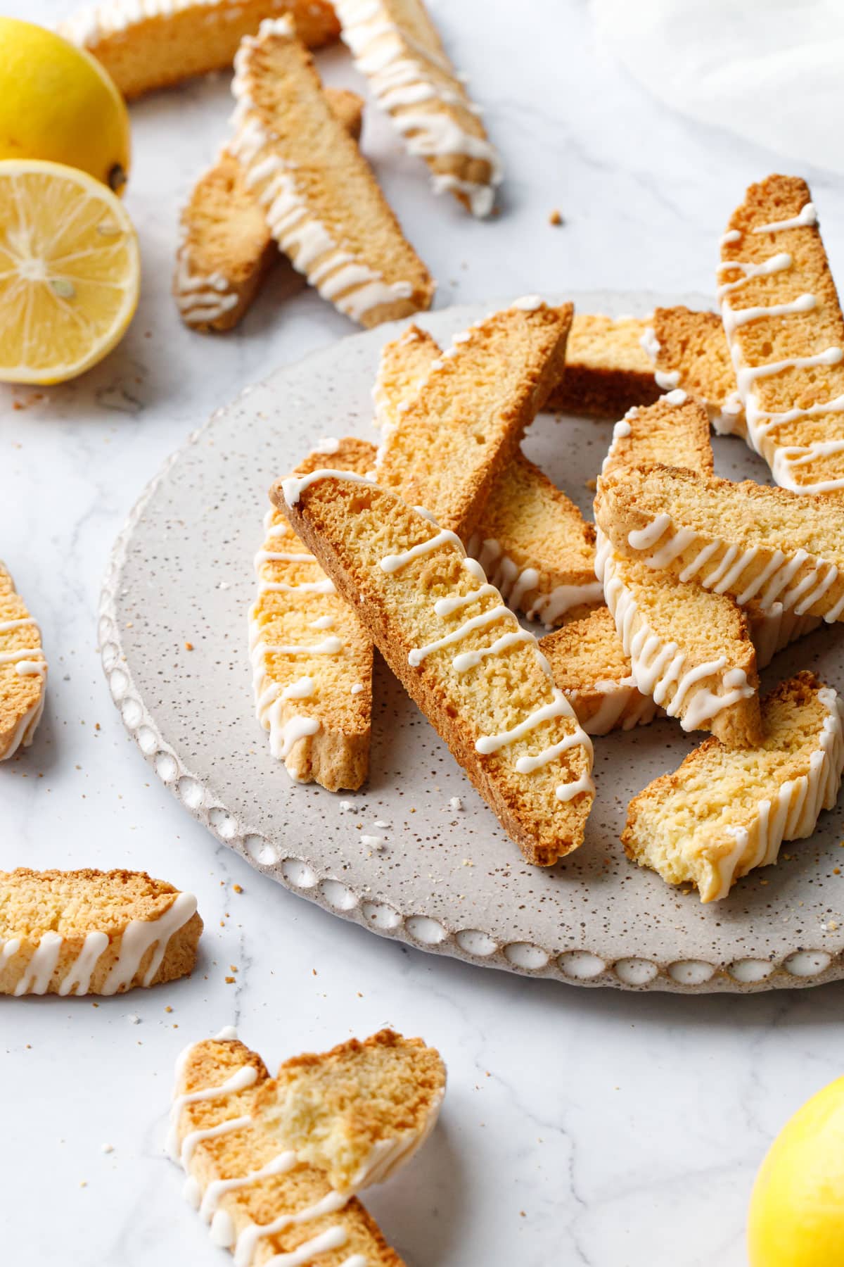 Scalloped ceramic cake plate piled with glazed Meyer Lemon Biscotti, more cookies scattered on the sides with a few fresh Meyer lemons too.