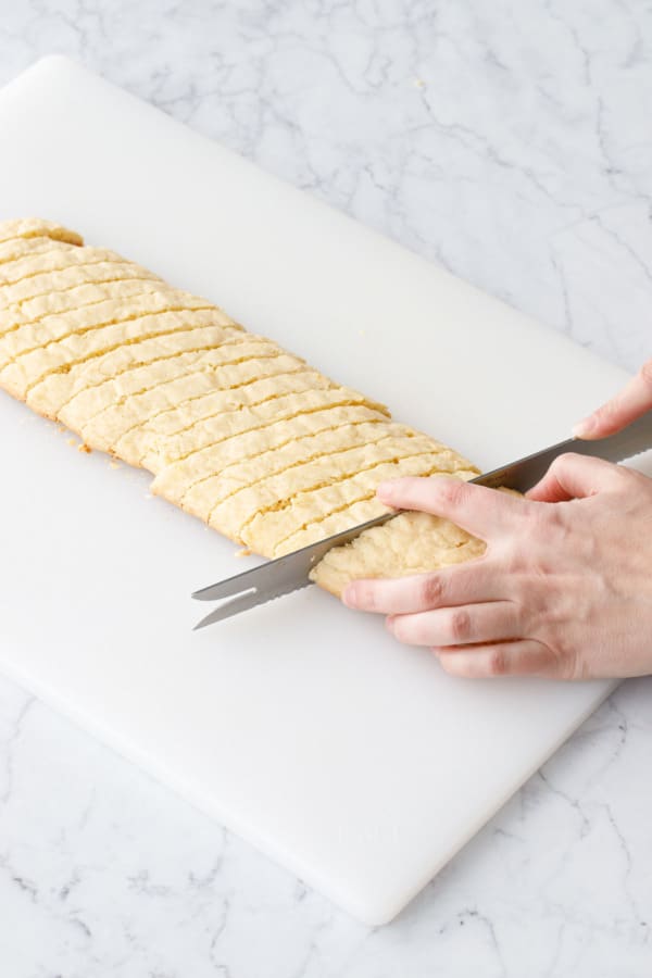 Serrated knife cutting diagonal slices of biscotti from the once-baked log.