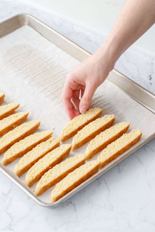 Laying cut slices of biscotti back on parchment-lined baking sheet in rows with very little space between cookies.