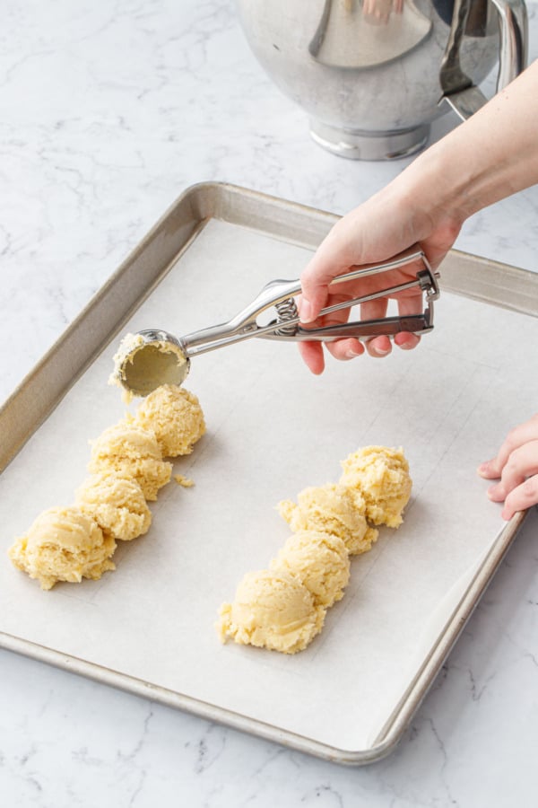 Cookie scoop dropping dollops of cookie dough into two rows on a parchment-lined cookie sheet.