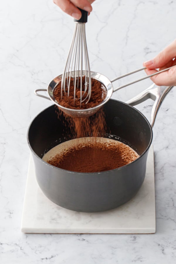 Sifting cocoa powder through a fine mesh sieve into a small saucepan filled with warm heavy cream.