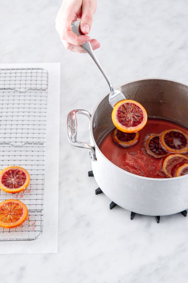 Fork lifting orange slice out of pot with sugar syrup.