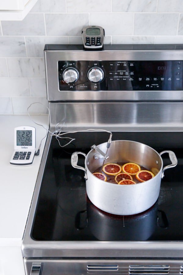 Stovetop with saucepan filled with sugar syrup and an even layer of blood orange slices, with a probe thermometer clipped on the side.
