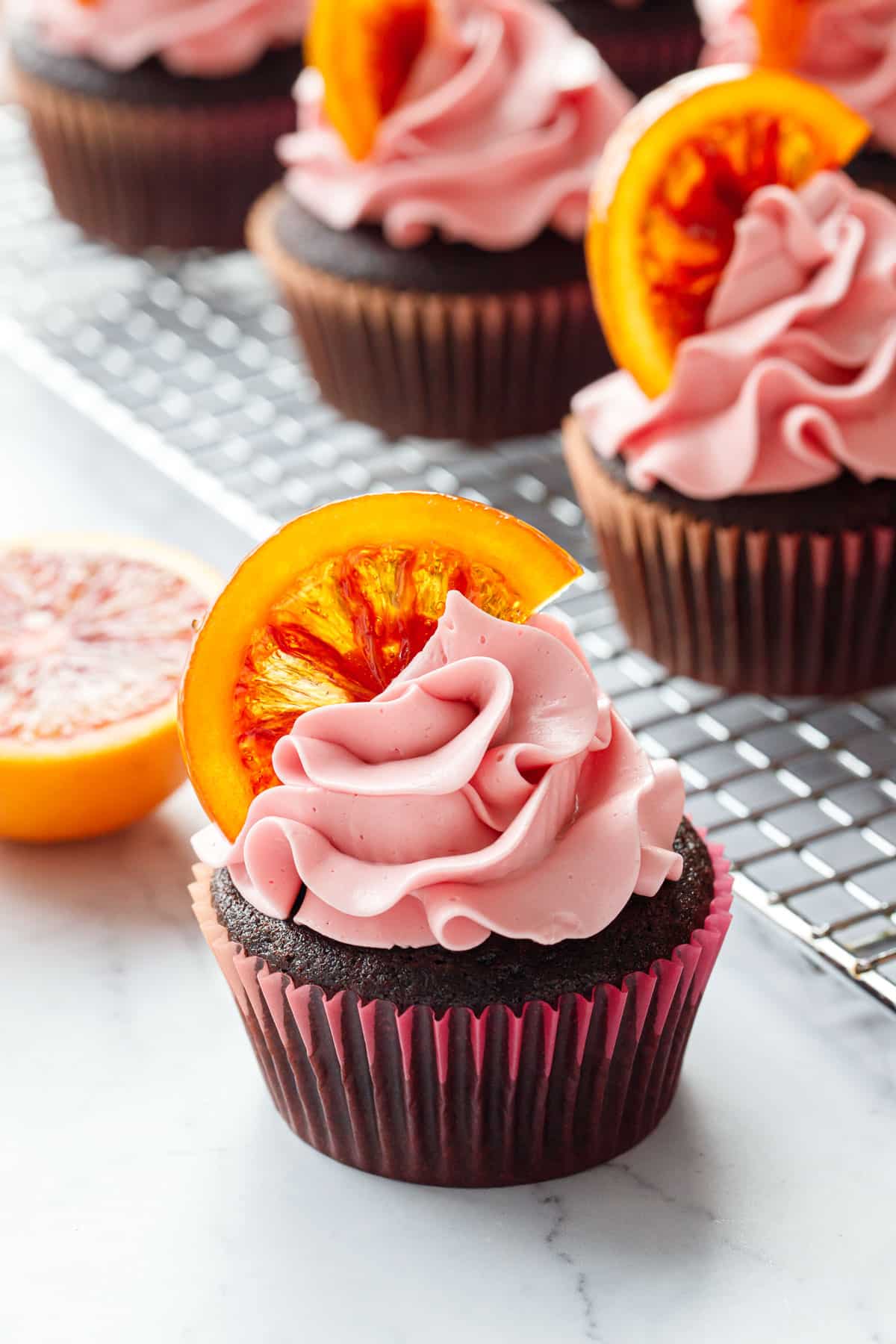 Closeup of Chocolate Olive Oil & Blood Orange Cupcake with a swirl of pink buttercream and topped with a candied blood orange slice, more cupcakes and half an orange in the background.