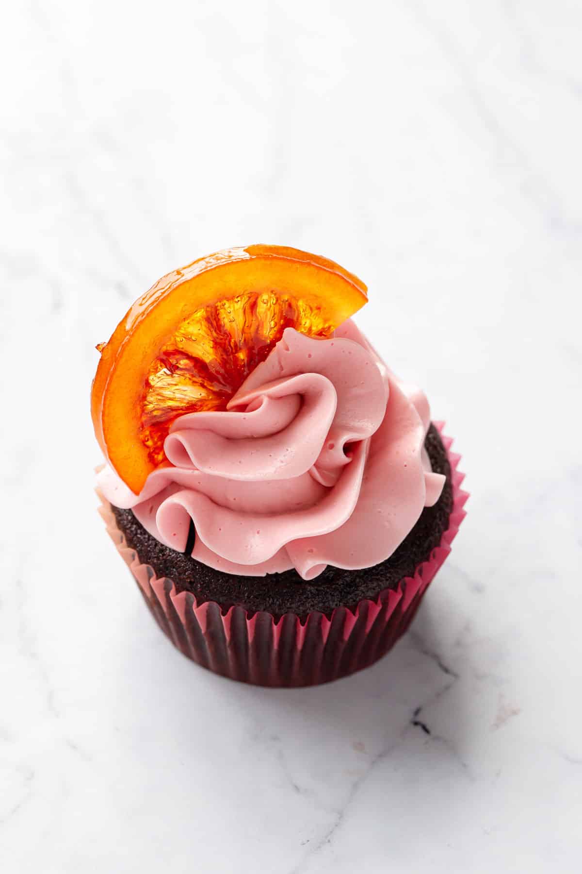 Closeup of a single Chocolate Olive Oil & Blood Orange Cupcake, topped with a swirl of light pink blood orange buttercream and a half moon candied blood orange slice.