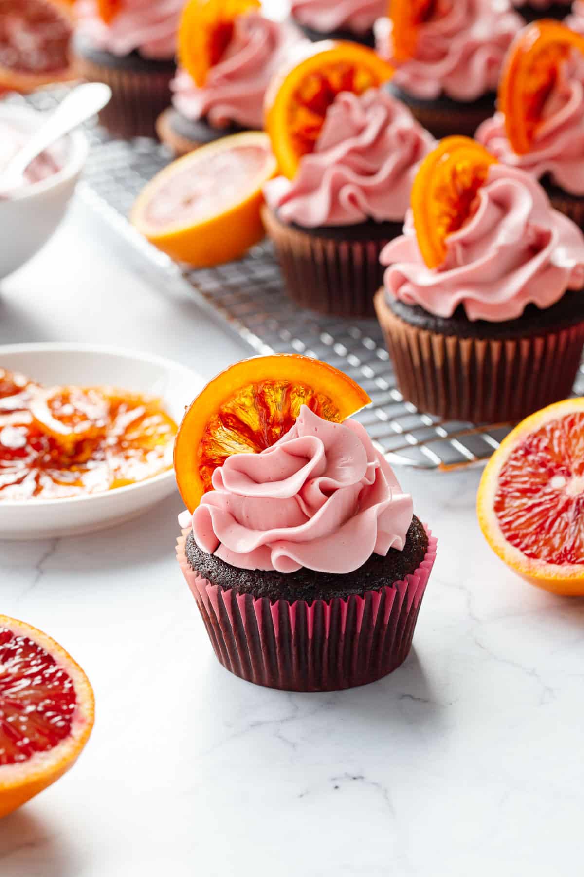 Chocolate Olive Oil & Blood Orange Cupcakes on a marble background, topped with swirls of pink frosting and halved slices of candied blood orange.