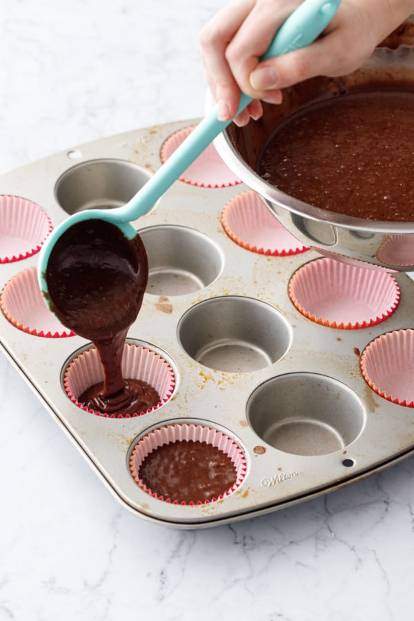Silicone ladle pouring chocolate cupcake batter into paper liners in a muffin tin.