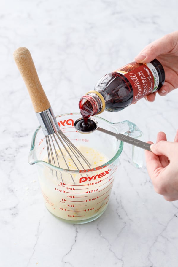 Measuring a teaspoon of Amoretti Natural Blood Orange Artisan Flavor above a measuring cup with other wet ingredients.