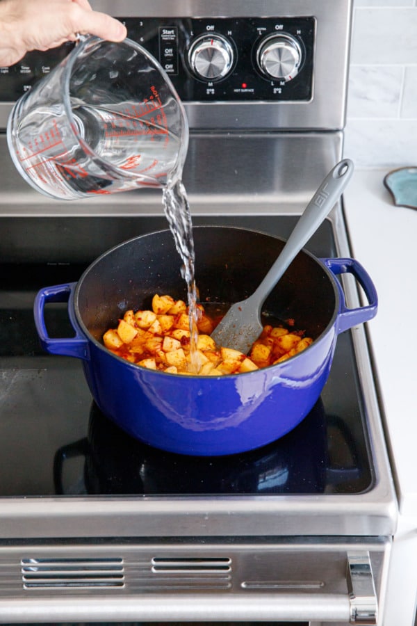 Pouring water into Dutch oven on stovetop, with tomato paste-covered potatoes inside.