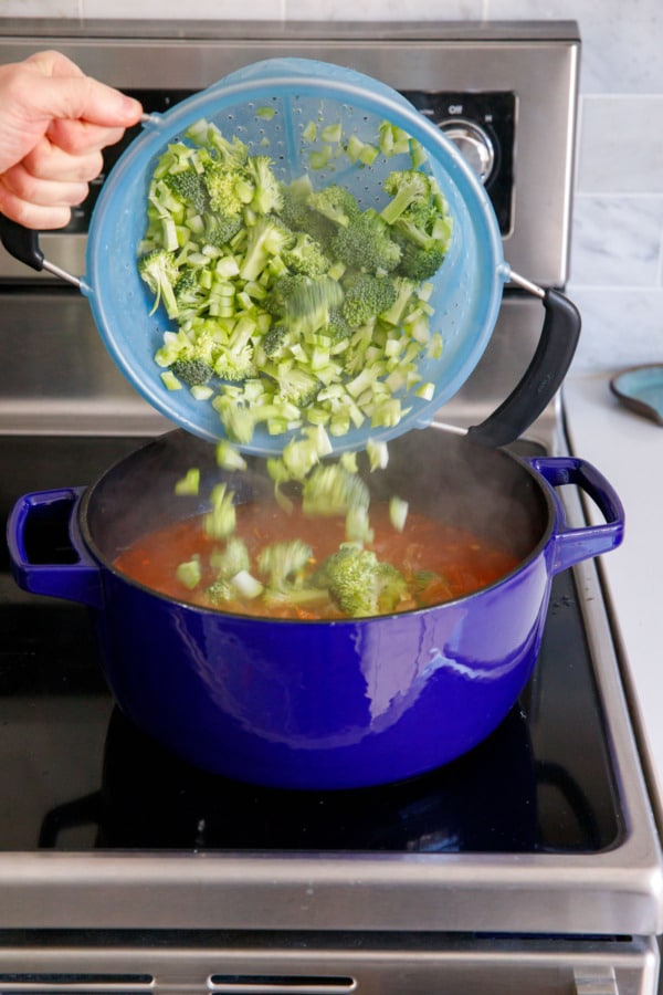 Dumping broccoli florets and chopped bits of stem into a Dutch oven filled with the tomato/potato mixture.