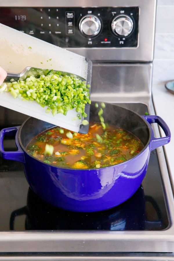 Scraping chopped green onions off a cutting board into a Dutch oven filled with broccoli & potato soup.