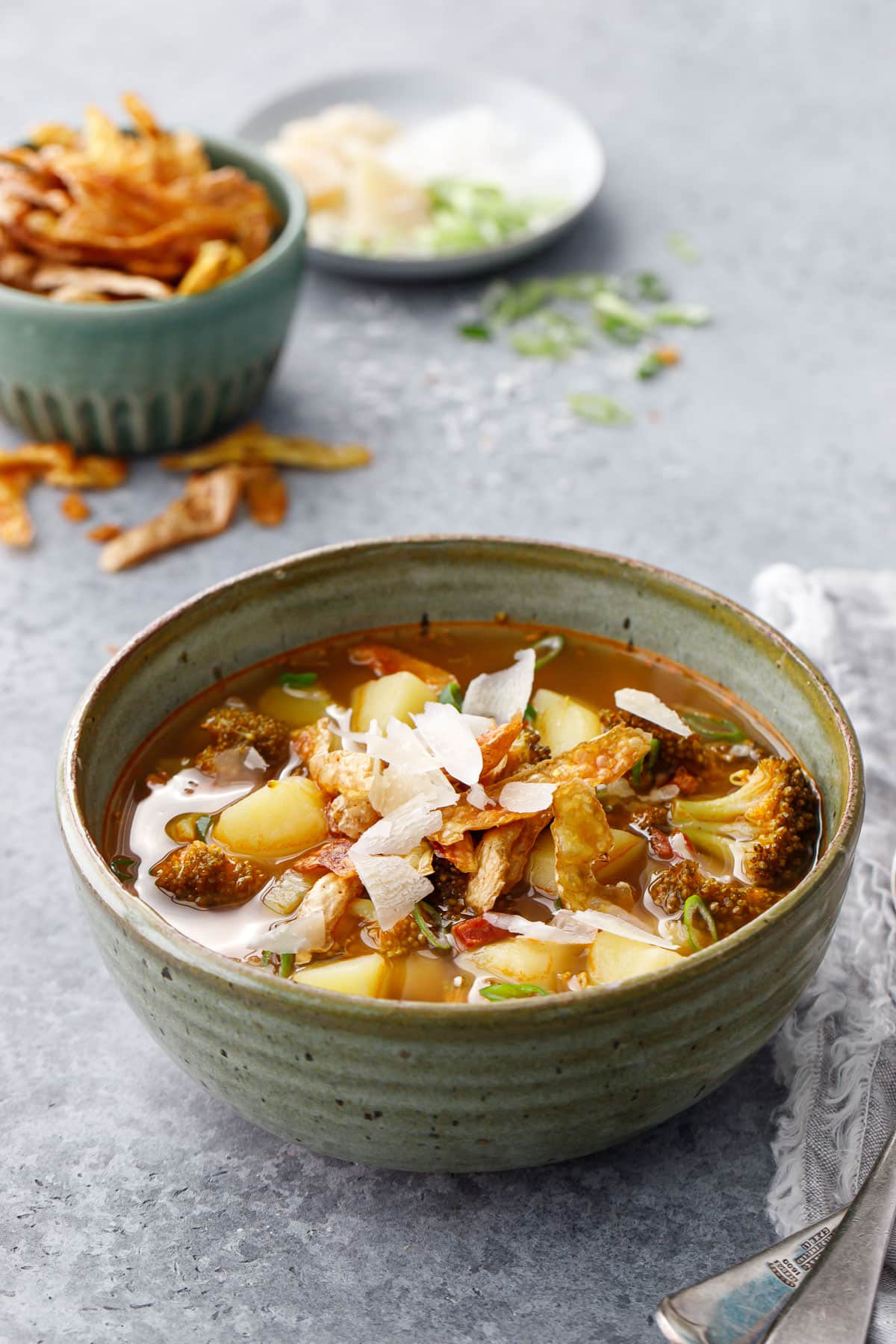 Brothy Broccoli & Potato Soup with Crispy Potato Skins in a ceramic bowl with napkin, with bowls of toppings in the background.
