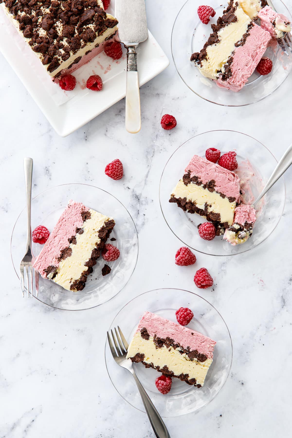 Overhead, marble background and three glass plates with cross-section slices of Raspberry & Passionfruit Semifreddo with Chocolate Crumbs, and a few frozen raspberries scattered around.