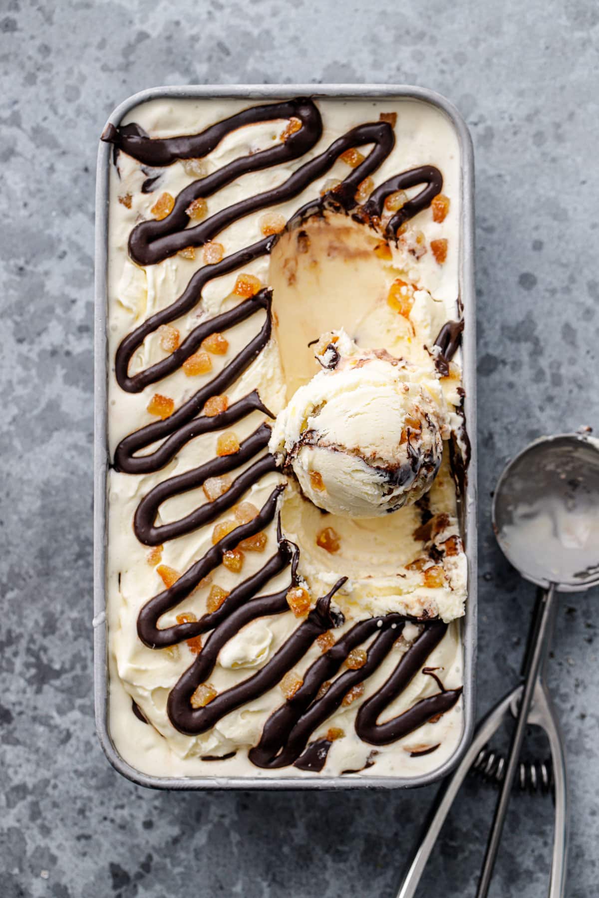 Overhead, silver loaf pan with candied orange ice cream topped with candied orange bits and a swirl of chocolate fudge, with one scoop scooped out.