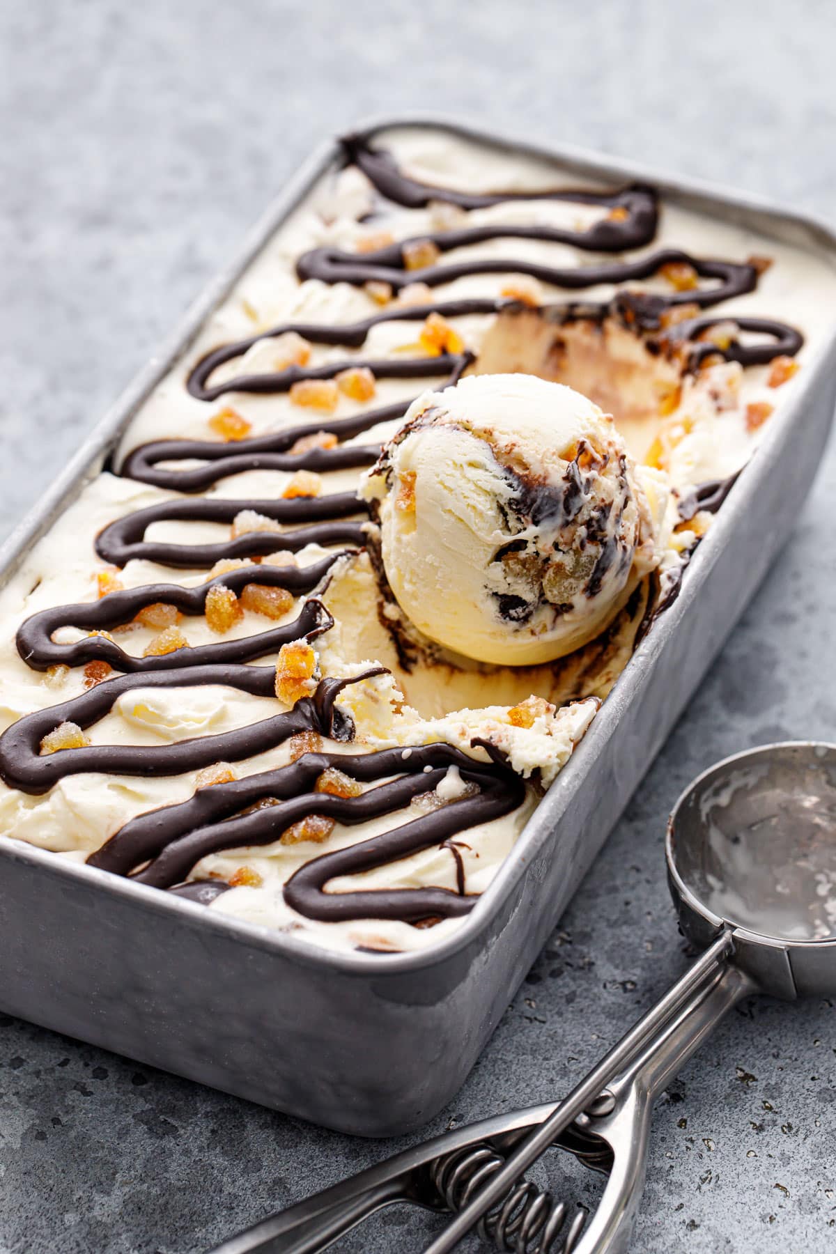 One round scoop sitting in a silver pan with Candied Orange Ice Cream with Chocolate Fudge Swirl, ice cream scoop on the side.