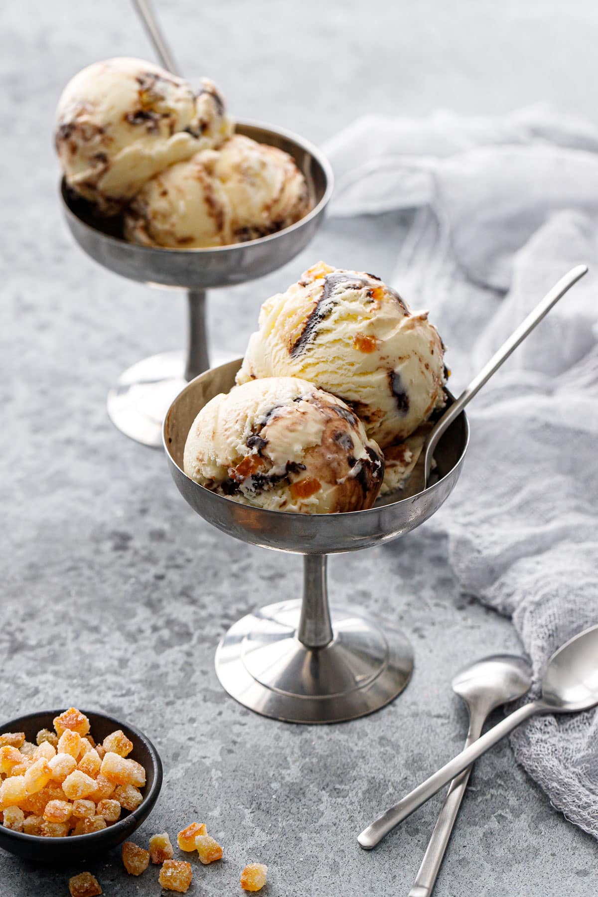 Two stainless sundae cups with scoops of Candied Orange Ice Cream with visible swirls of chocolate fudge, small bowl of candied oranges and two spoons on the side.