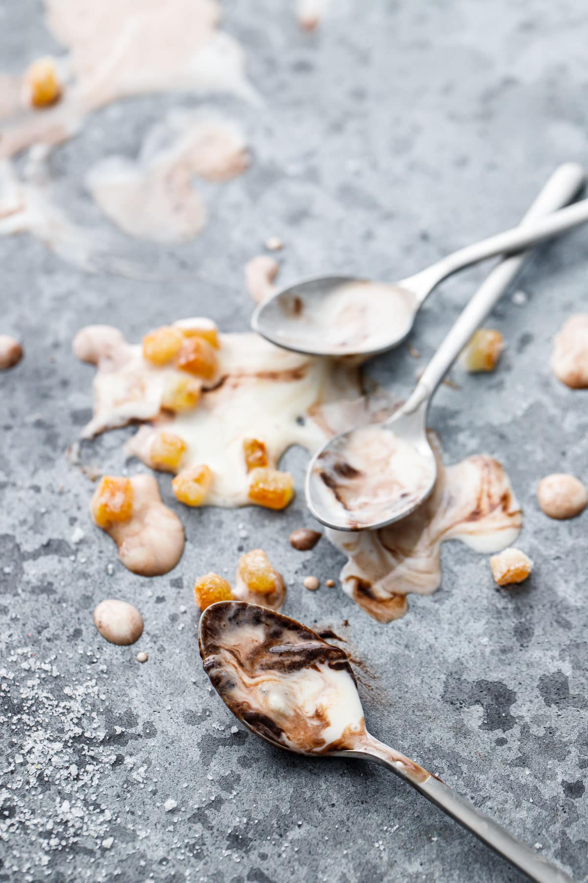 Messy scene with two spoons and melted ice cream swirled with fudge and candied orange bits on a gray background.