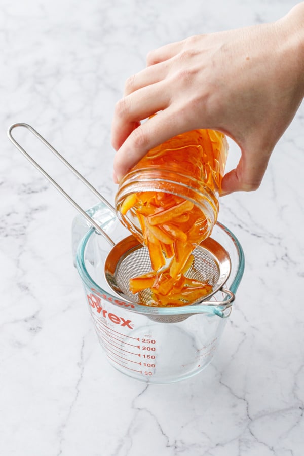 Pouring a jar of candied orange peel and syrup through a fine mesh sieve to strain the syrup.