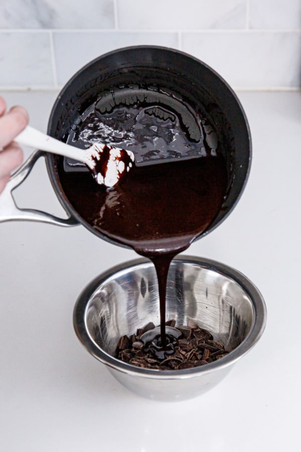 Pouring hot fudge sauce over chopped dark chocolate in a stainless steel bowl.