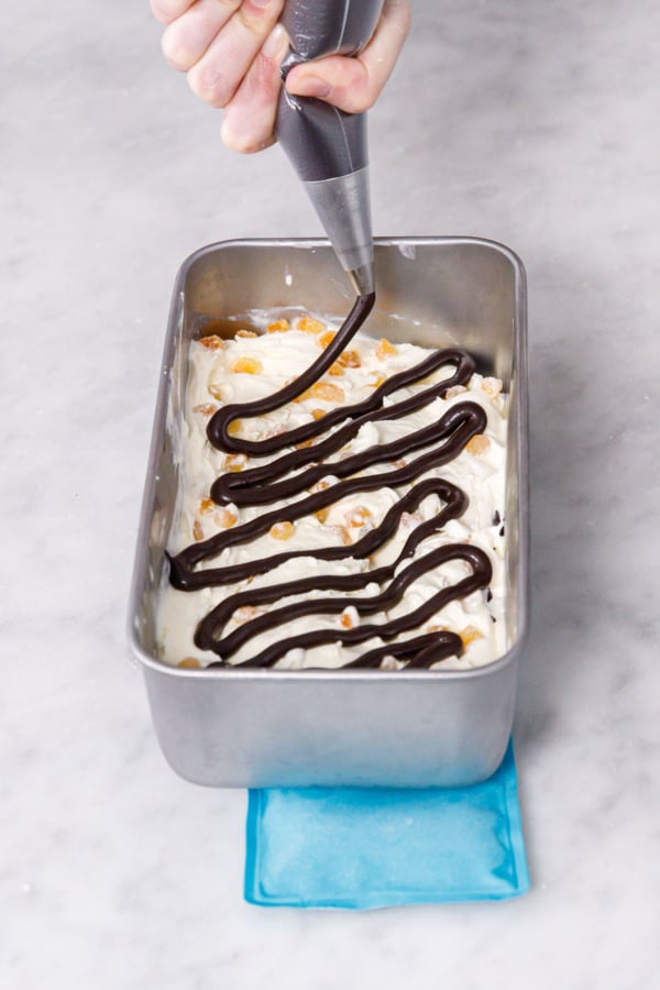 Piping bag drizzling a thick squiggle of fudge sauce on top of churned ice cream in a loaf pan.