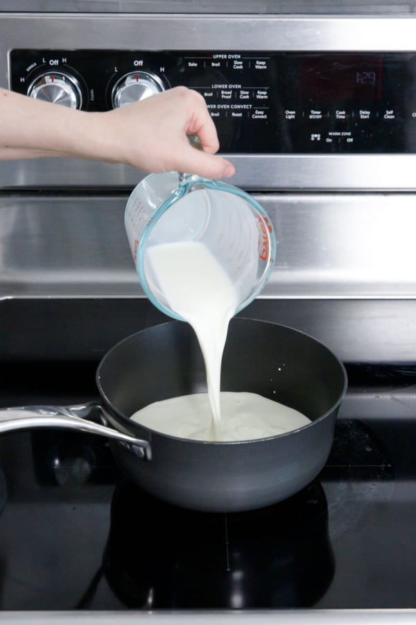 Pouring heavy cream and milk into a saucepan on a glass cooktop.