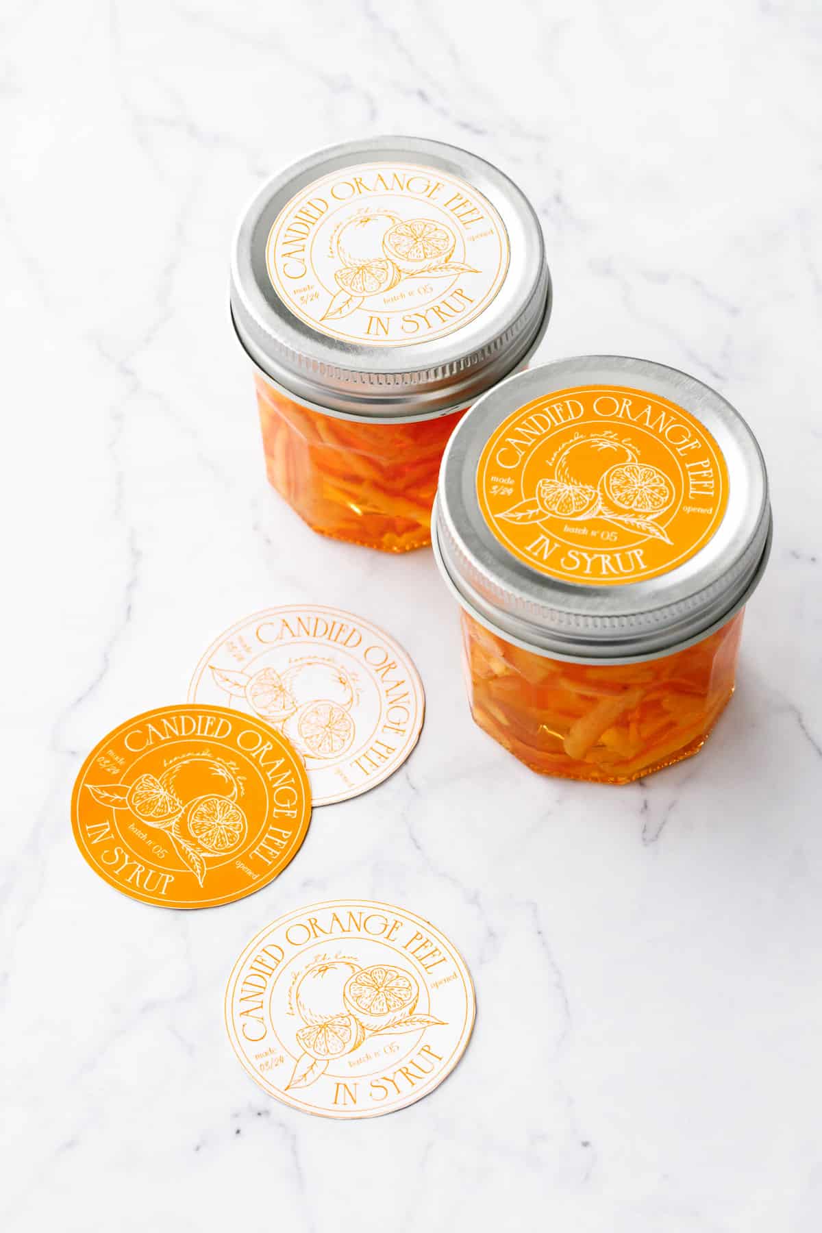 Jars of Candied Orange Peel in Syrup with pretty designed orange and white labels, and a few more circular cut out labels sitting on the side.
