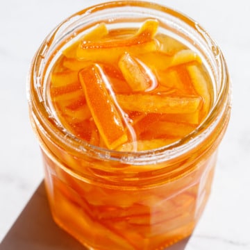 Direct light on an open glass canning jar of Candied Orange Peel in Syrup on a marble background.