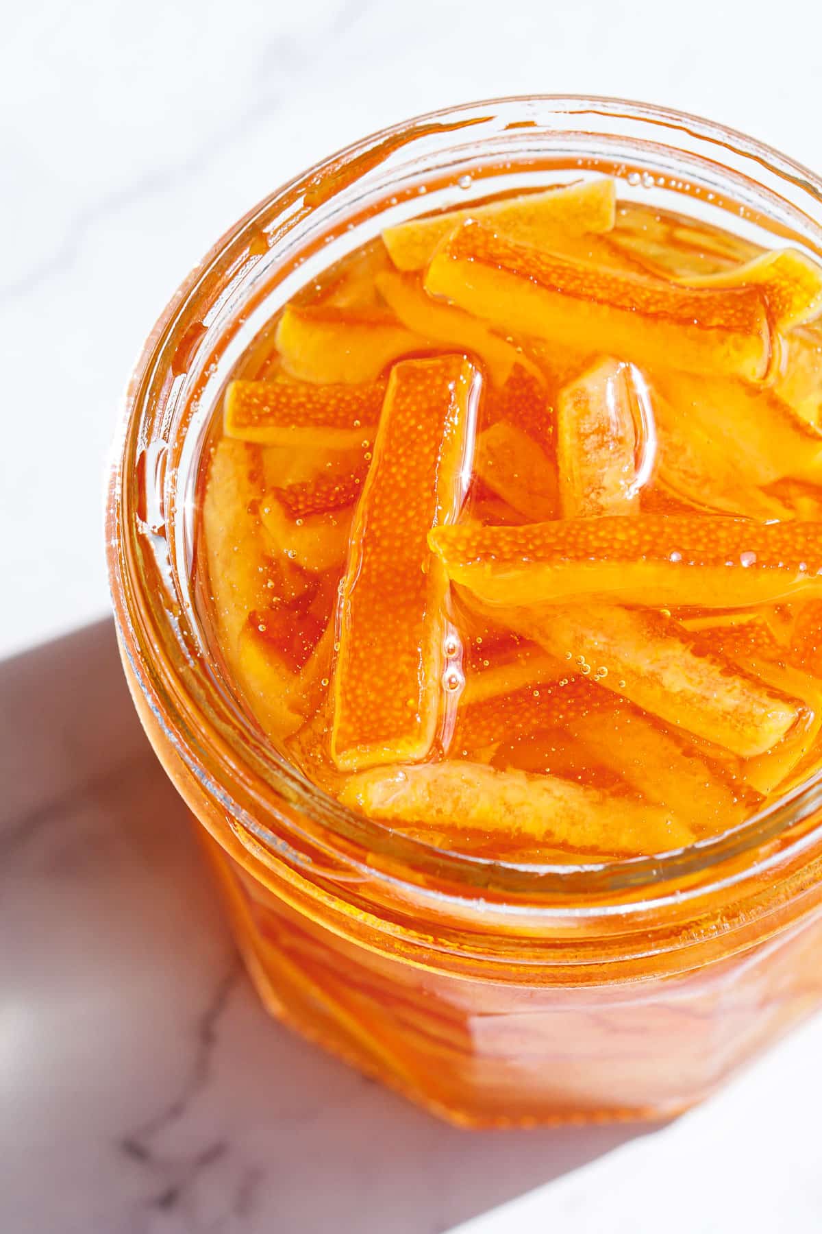 Closeup, open glass canning jar filled with Candied Orange Peel in Syrup, showing the texture of the candied orange peel and crystal clear sugar syrup in direct sunlight.