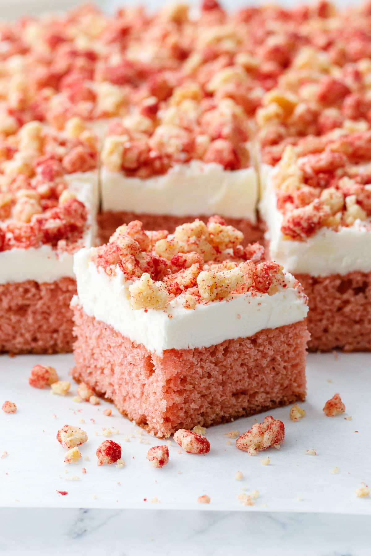 Closeup cut square of Strawberry Crunch Sheet Cake with Buttermilk Frosting showing the light pink crumb and thick layer of fluffy white buttercream with messy crumbs scattered around.